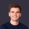Portrait of a young man with short light brown hair and a trimmed beard, smiling slightly, wearing a black shirt, against a dark blue background.
