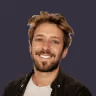 Smiling young man with light brown hair and beard wearing a black jacket on a dark background.