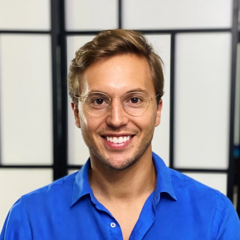 Smiling man with light brown hair wearing glasses and a bright blue collared shirt, standing in front of a blurred grid-patterned background.