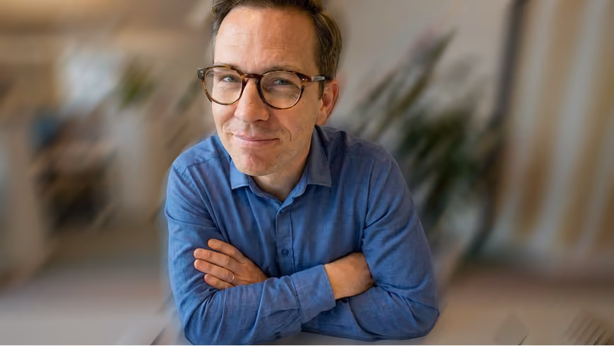 Smiling man wearing glasses and a blue shirt with arms crossed, looking at the camera in a blurred indoor setting.