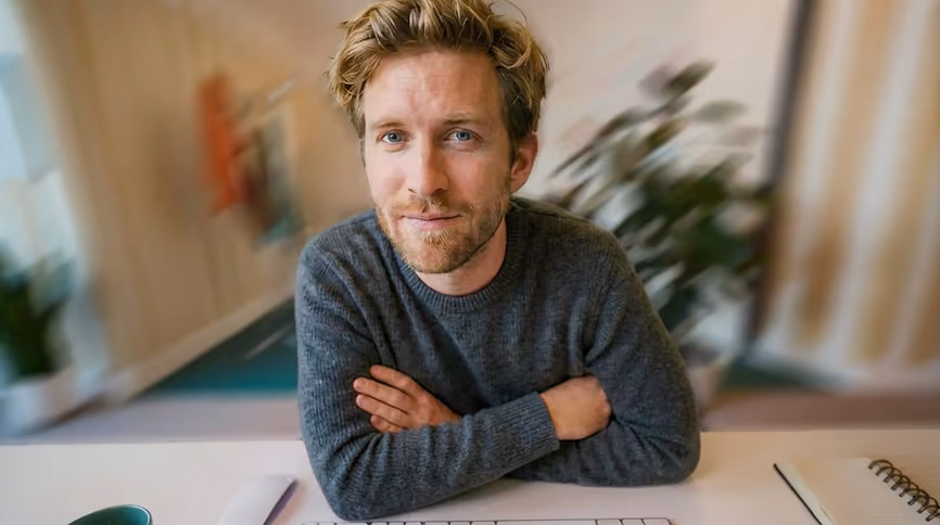 A man with blond hair and blue eyes wearing a gray sweater sits at a desk with arms crossed, looking directly at the camera, with a blurred background including plants and office items.