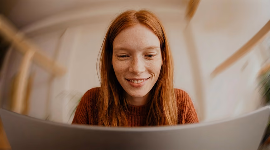 Close-up of a smiling woman with long red hair and freckles looking at a laptop screen in a warmly lit room.