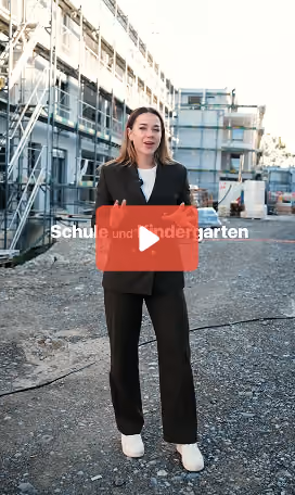Eine Frau in einem schwarzen Kostüm und weißen Schuhen steht auf einem Schotterweg vor einem im Bau befindlichen Gebäude mit Gerüsten und spricht in die Kamera.
