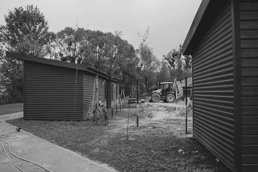 Black and white image showing a row of small corrugated metal buildings with pitched roofs along a paved path, young trees supported by stakes in grassy soil, and a tractor in the background near a puddle of water.