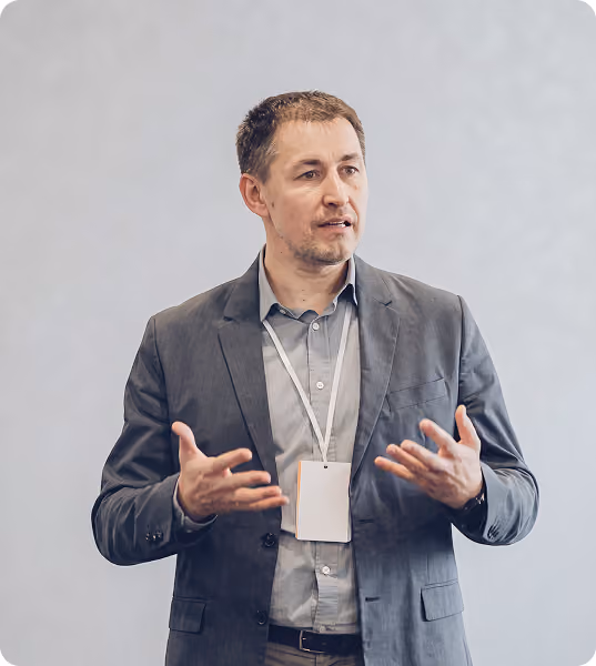 Man in a gray blazer and light gray shirt wearing a name badge on a white lanyard, speaking with hands gesturing, against a plain light gray background.
