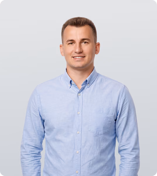 Smiling man with short brown hair wearing a light blue button-up shirt standing against a plain light gray background.
