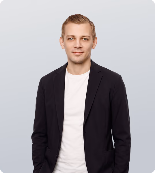 A man with short light brown hair wearing a black blazer over a white shirt standing against a gradient light gray background.