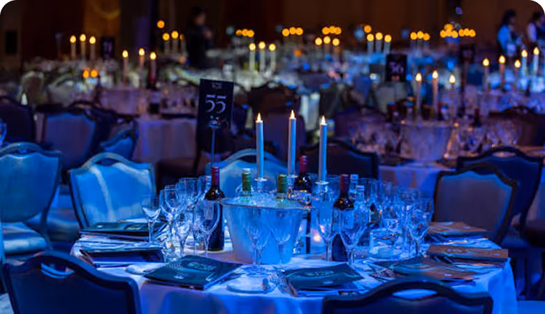 Round table set for a formal event with lit candles, wine bottles, multiple glasses, folded napkins, and menus under dim, blue ambient lighting.