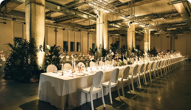 Long banquet table set elegantly with white chairs, candles, floral centerpieces, and greenery in an industrial-style room with exposed beams and concrete pillars.