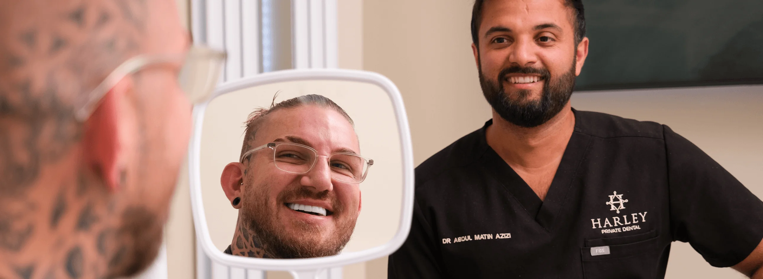 Smiling male patient with tattoos looking at his reflection in a dental mirror while a male dentist in black scrubs stands nearby.