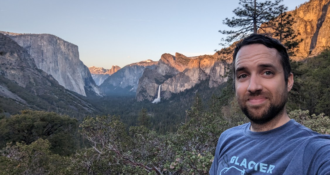 A man with short dark hair and a beard wearing a blue Glacier National Park T-shirt takes a selfie with a scenic view of Yosemite Valley in the background, featuring steep cliffs, a waterfall, pine trees, and a clear sky during sunset.