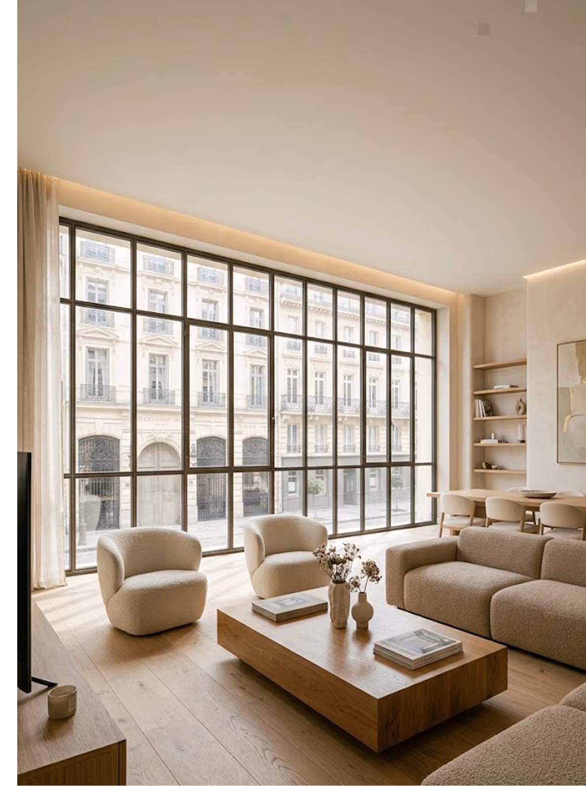 Séjour contemporain rénové par un architecte d'intérieur, avec une grande verrière à cadre noir donnant sur une façade ancienne, canapé et fauteuils bouclette crème, table basse en bois massif, parquet en chêne clair et éclairage indirect