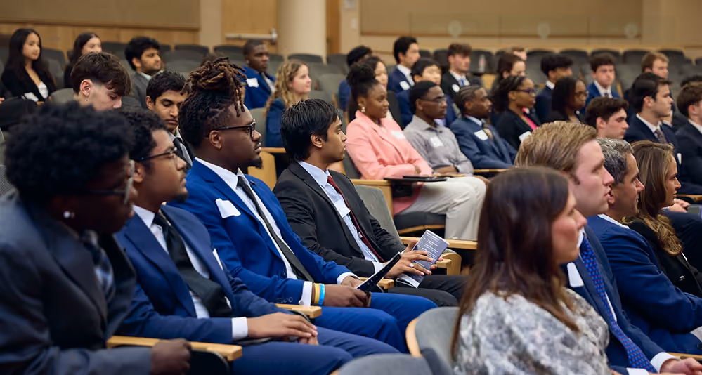 Students in business attire seated in an auditorium, several reading event programs