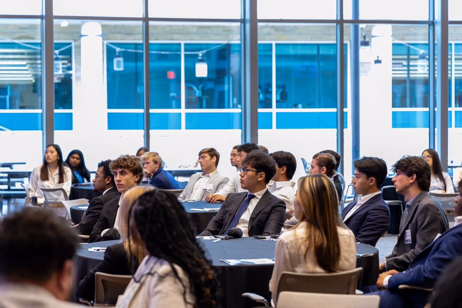 Students seated at round tables in a bright conference room watching a presentation
