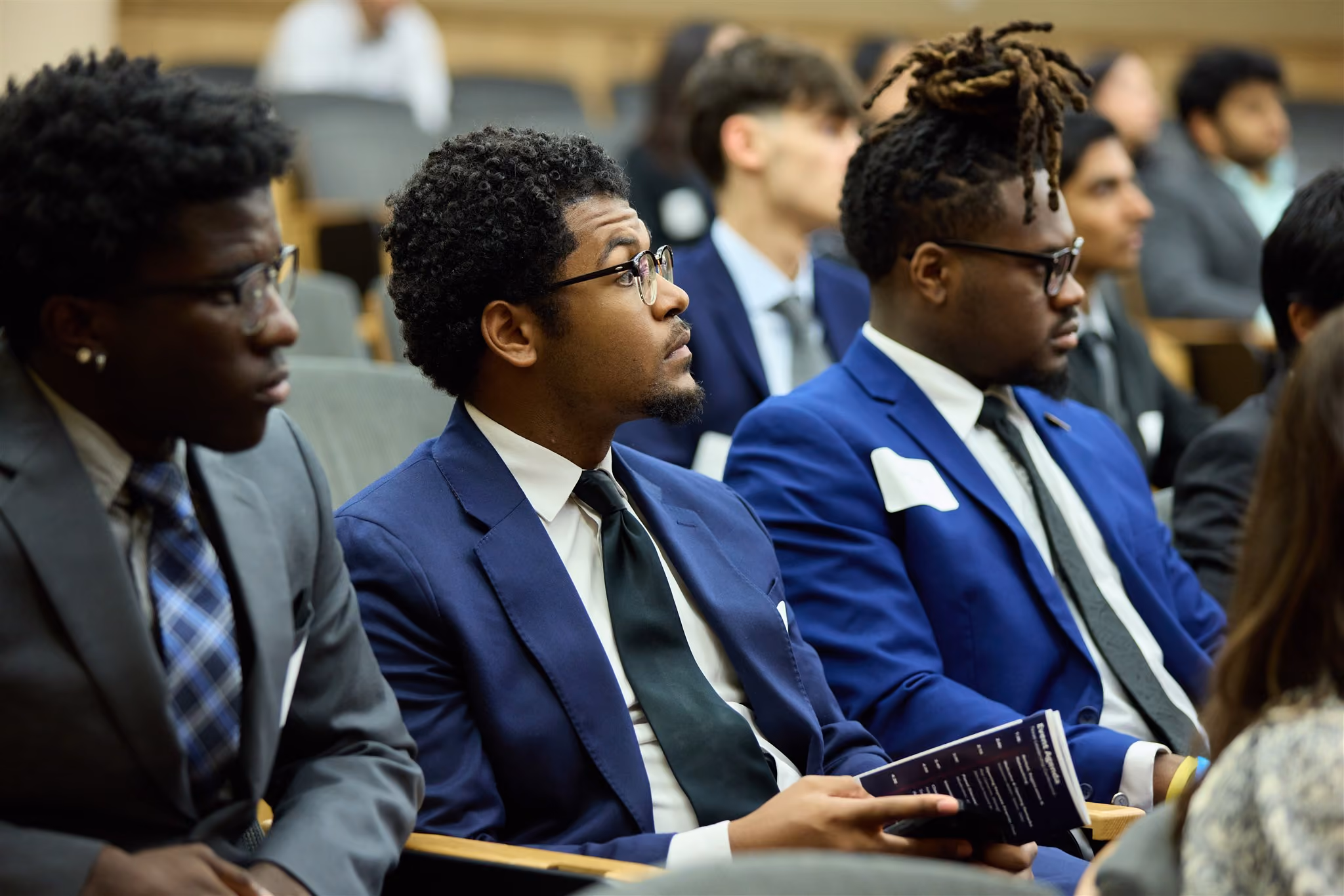 Students in suits seated in an auditorium listening to a presentation, one holding a program