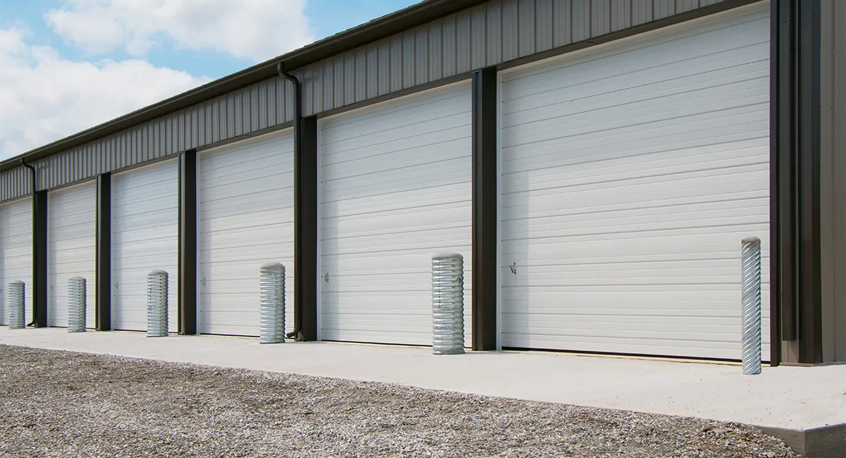 Row of white sectional garage doors on a commercial building with a concrete walkway and metal bollards in front under a partly cloudy sky.