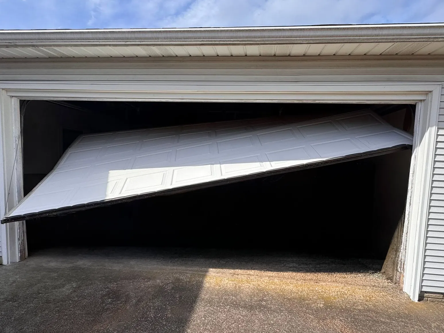 White garage door partially detached and hanging unevenly at an open garage entrance with concrete driveway in front.