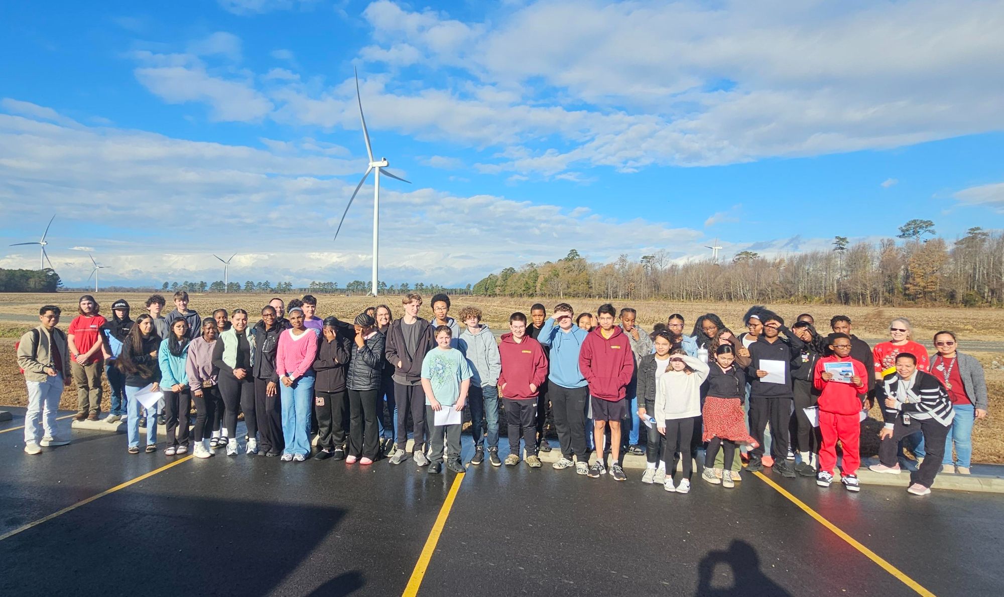 Group of diverse students and teachers standing outside near wind turbines on a sunny day.