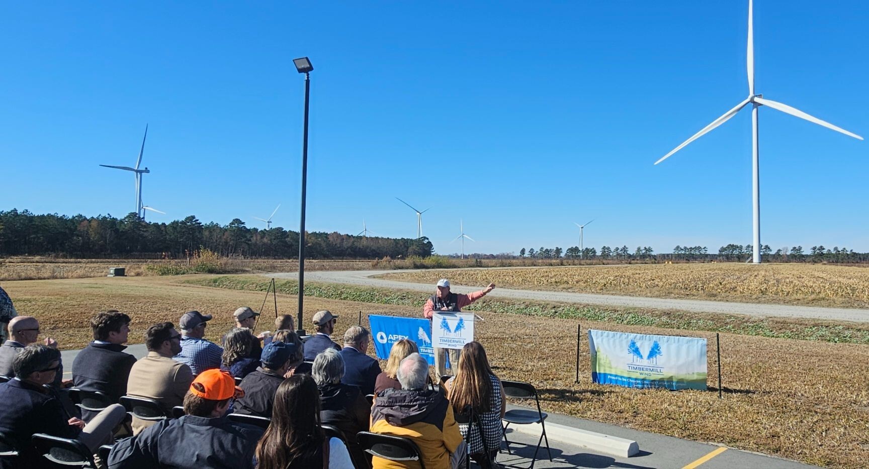 Speaker at outdoor event with wind turbines and seated audience under clear blue sky.