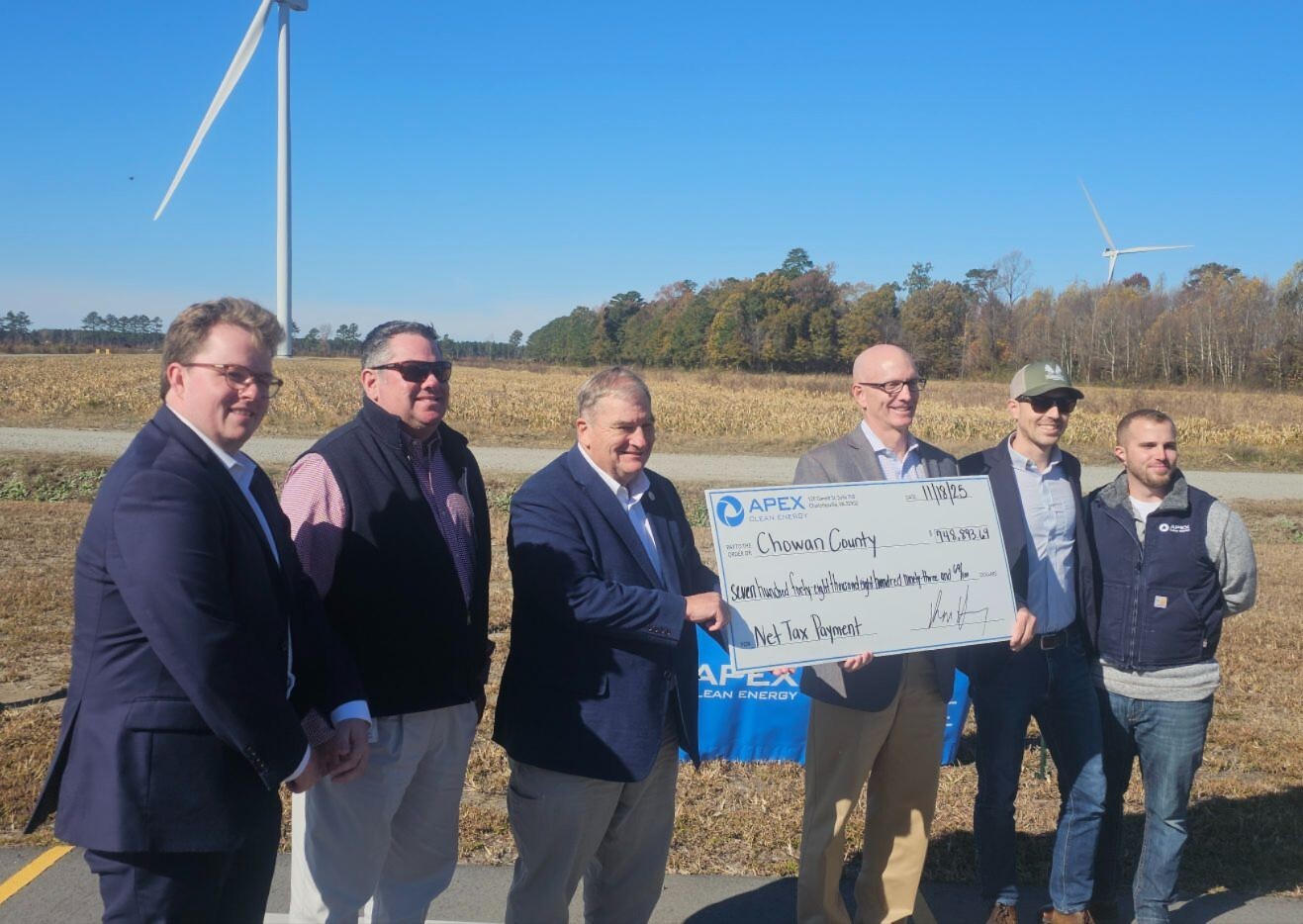 Six men standing outdoors in front of a field with wind turbines, holding a large check from APEX Clean Energy made out to Chowan County for $749,883.19 dated 11/18/25.