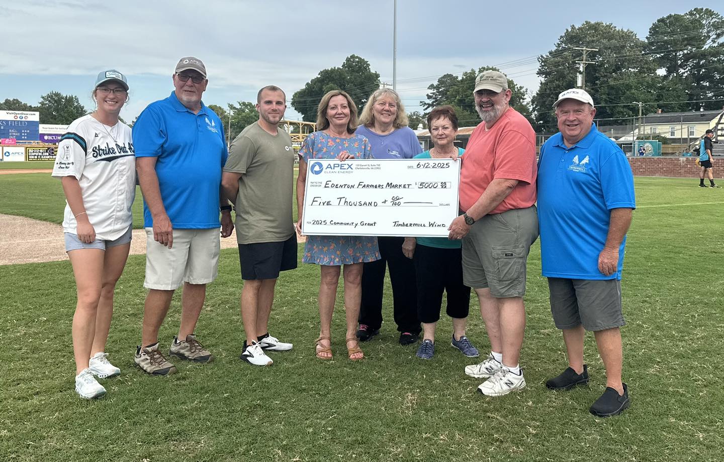 Eight people standing on a baseball field holding a large check for five thousand dollars made out to Edenton Farmers Market, dated 6-12-2025, from Apex Clean Energy and Timbermill Winds.