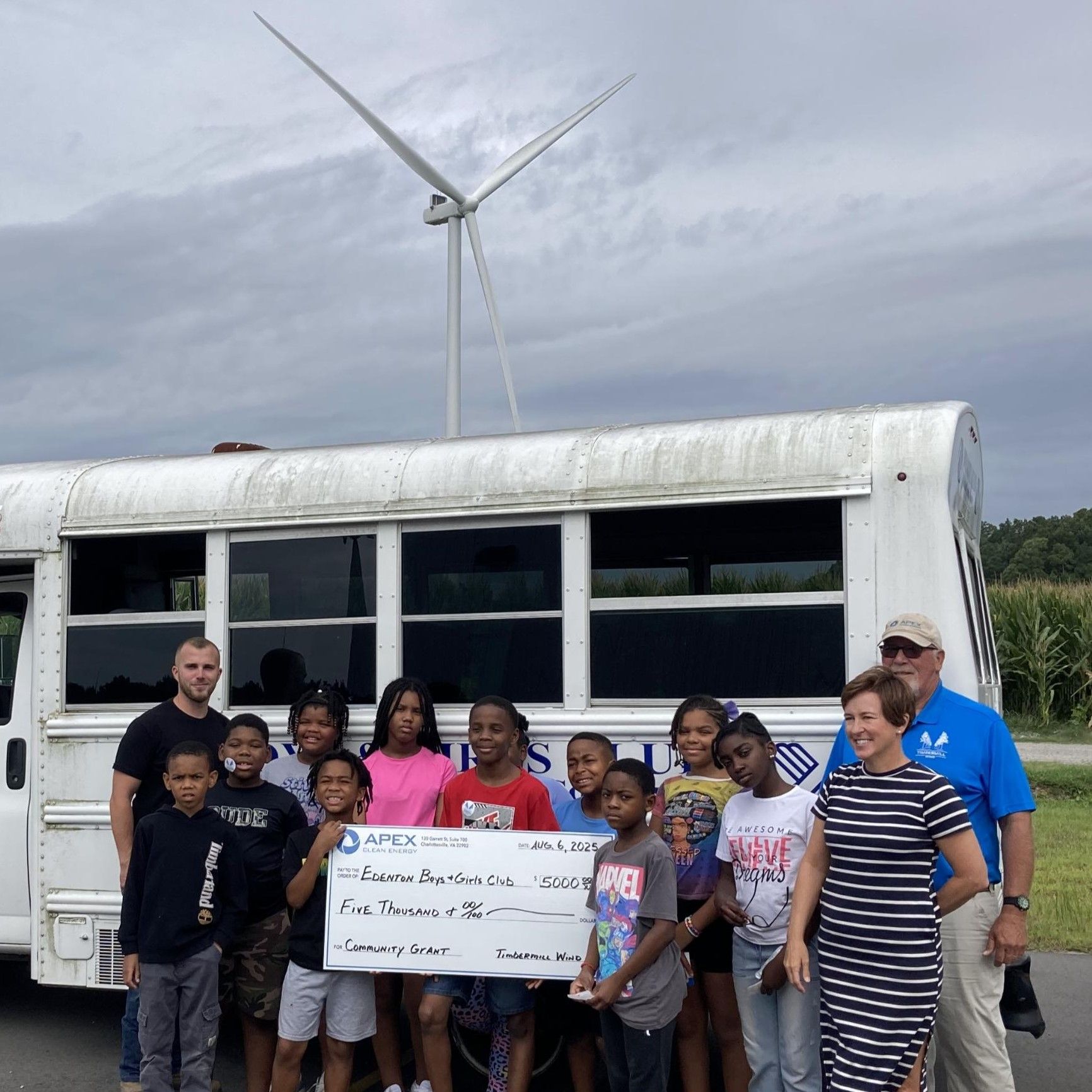 Group of children and adults holding a $5,000 community grant check in front of a bus with wind turbine behind.