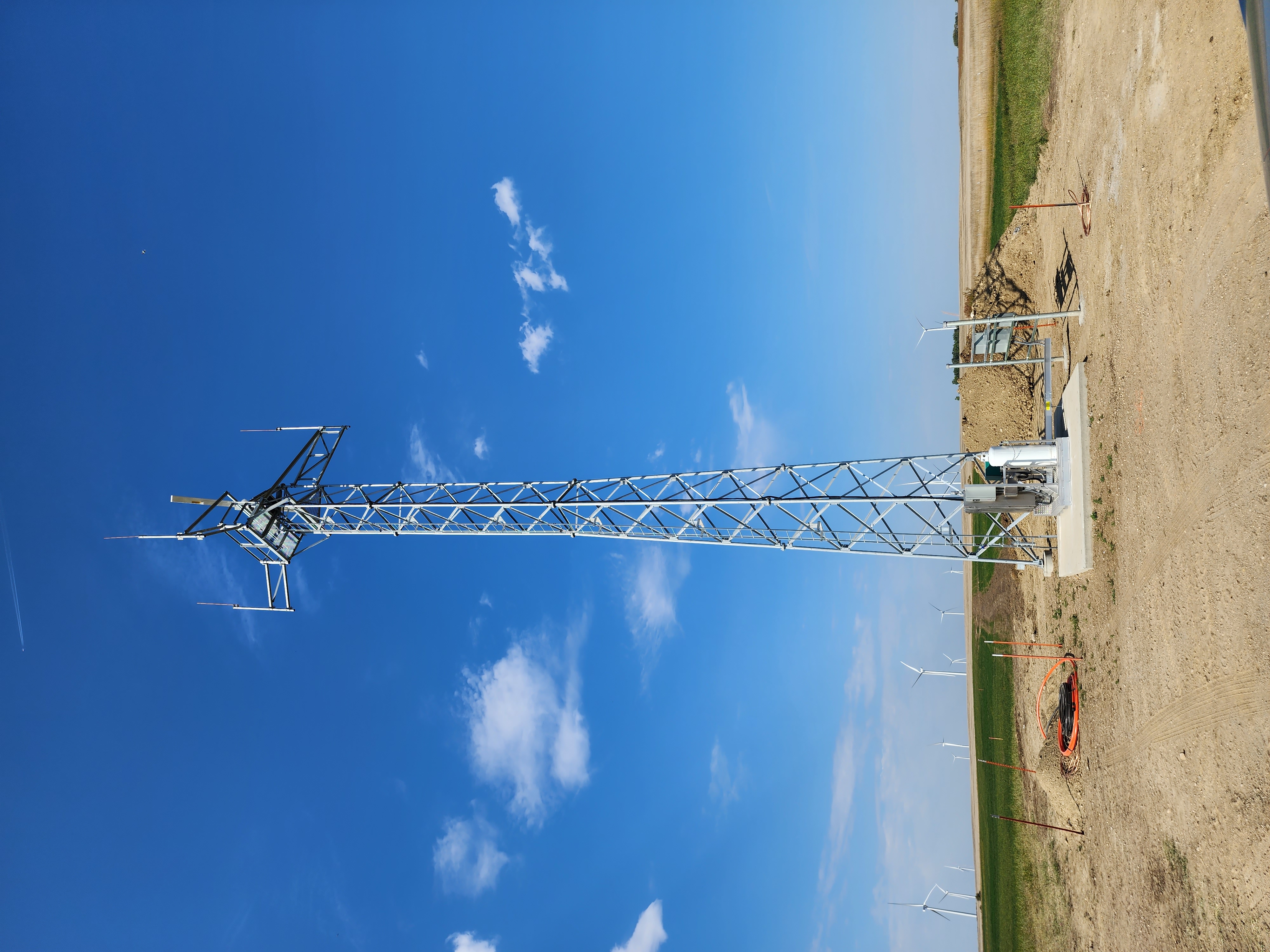 Metal communication tower stands on dirt land with wind turbines in the background under a blue sky.