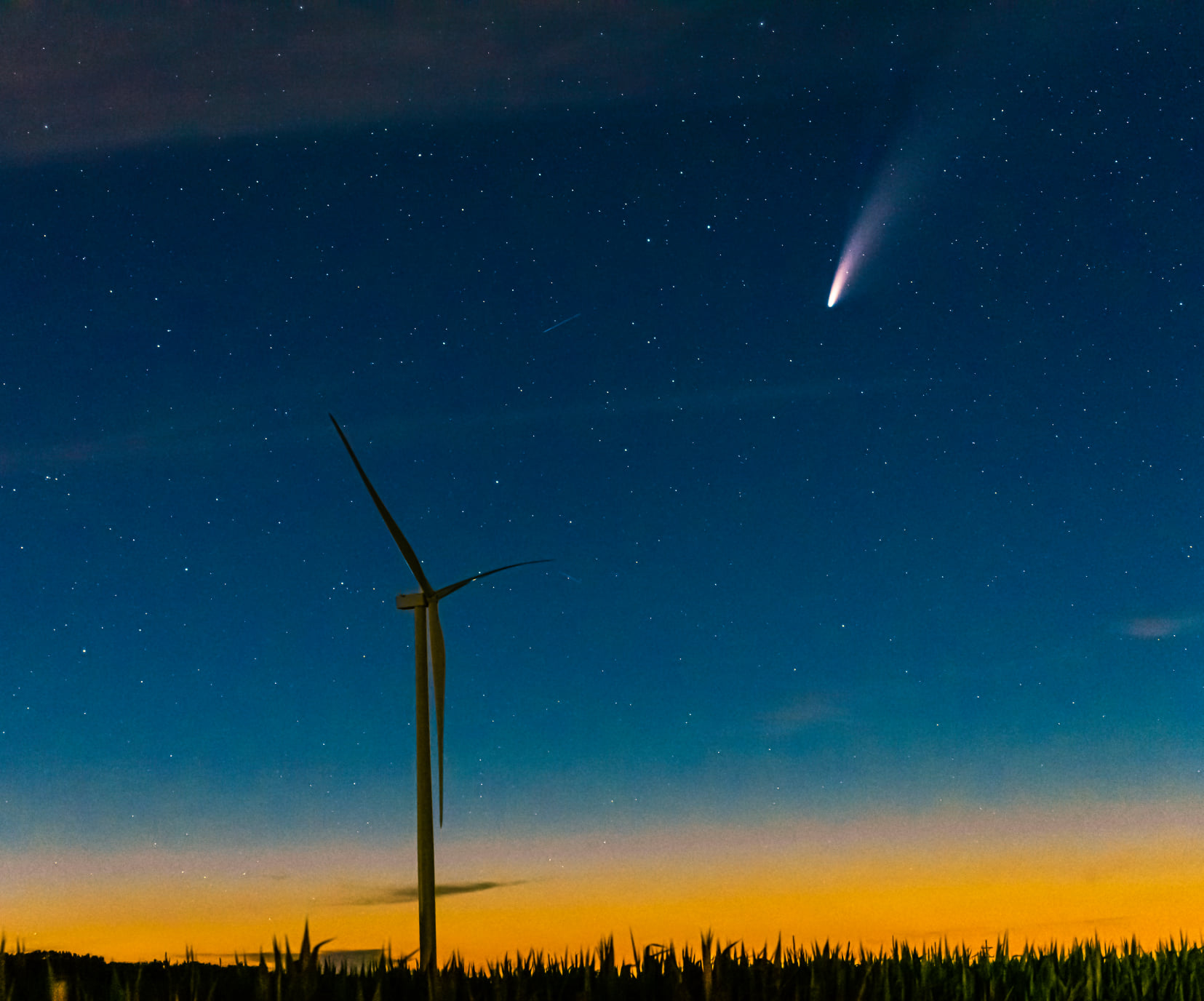 A wind turbine stands above a field at dusk with an orange horizon, under a clear night sky filled with stars and a bright comet with a glowing tail.