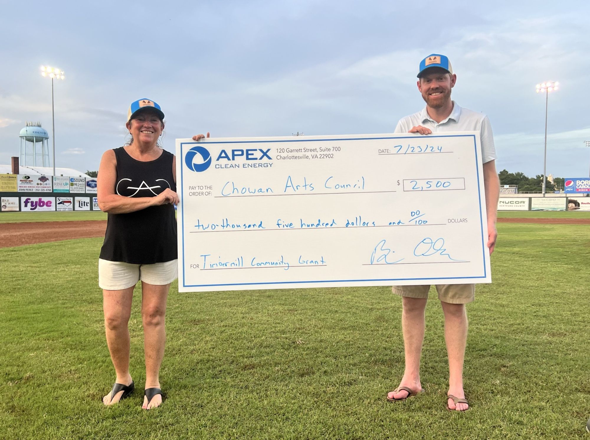 Two people standing on a baseball field holding a large check from Apex Clean Energy for $2,500 to Chowan Arts Council dated 7/23/24 for the Tidermill Community Grant.