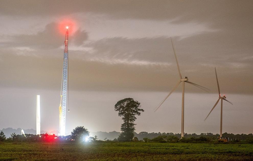 Nighttime scene of a field with two large wind turbines, one with red warning lights on the blades, and a tall construction crane with a red light at the top and bright white lights at the base, a tree is visible between the crane and turbines under a cloudy sky.