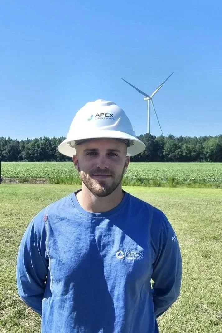 A man wearing a white hard hat and blue long-sleeve shirt with 'APEX Clean Energy' logos stands outdoors in front of a field and trees with a wind turbine in the background under a clear blue sky.