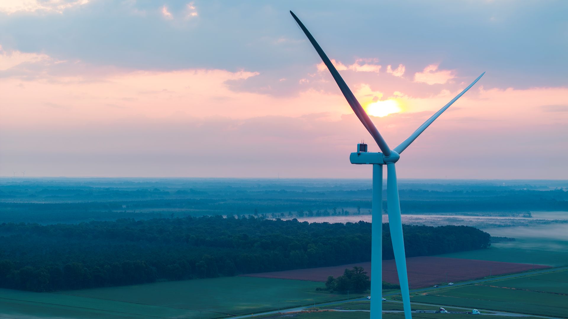 Close-up of a large wind turbine with two visible blades against a sunrise sky over a green forest and farmland landscape.