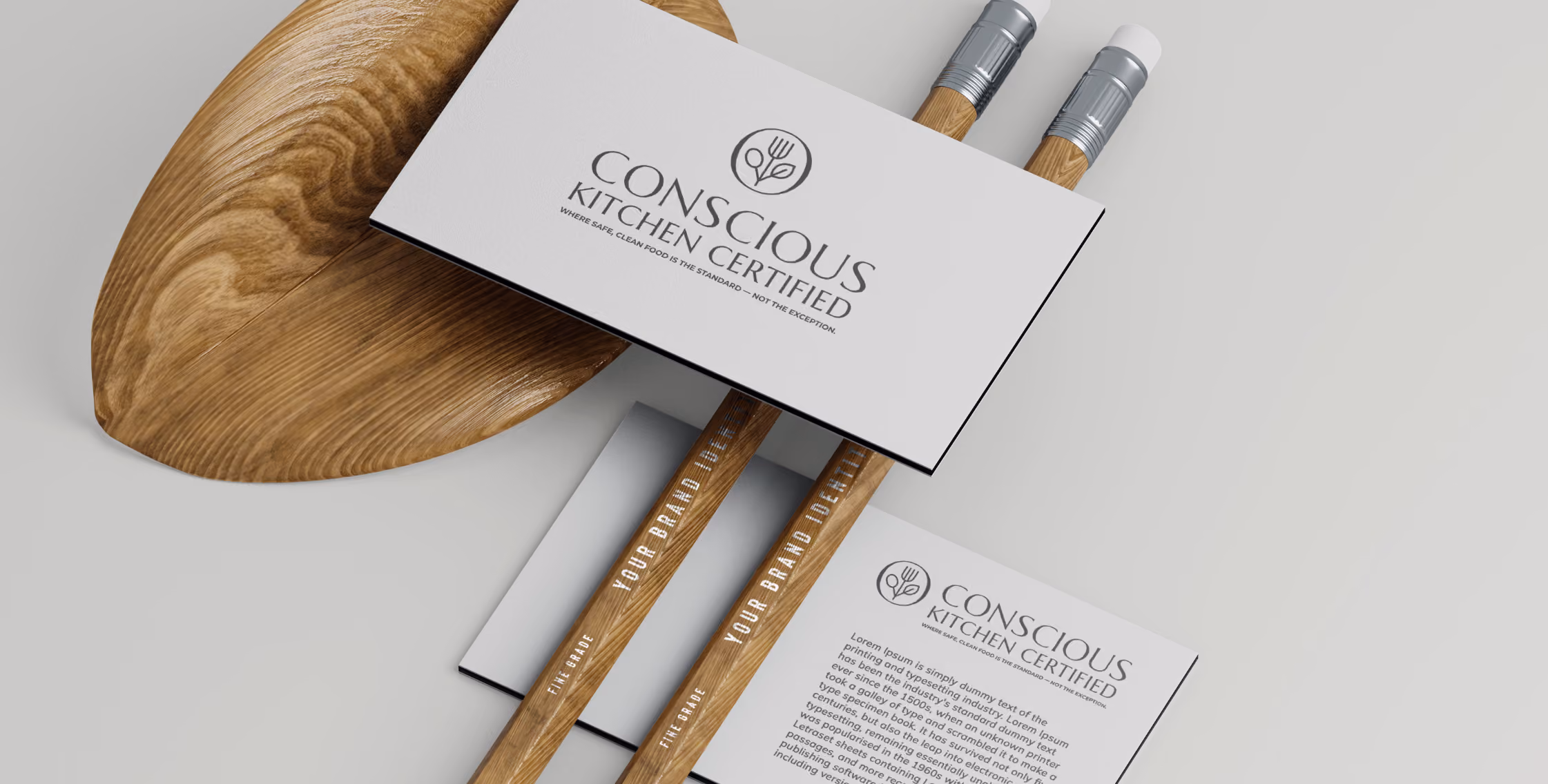 Two wooden pencils and business cards with the Conscious Kitchen Certified logo and slogan on a white surface, accompanied by a wooden cutting board.