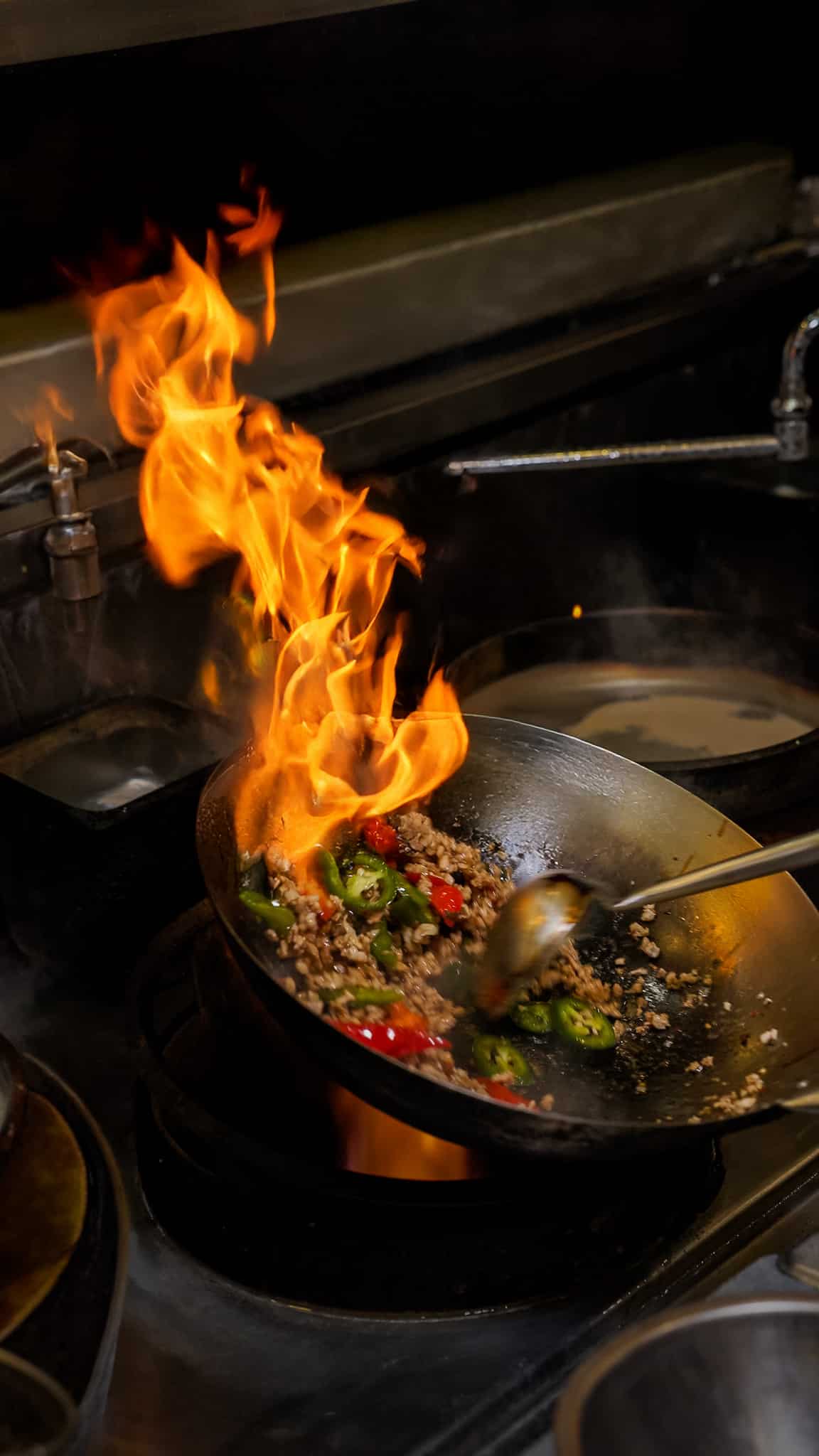 Flames leaping from a wok of minced meat and green chilies on a restaurant stove