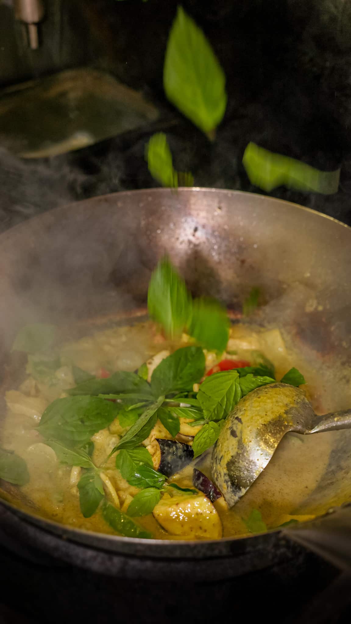 Green curry simmering in a steel wok with fresh basil leaves and eggplant, steam rising