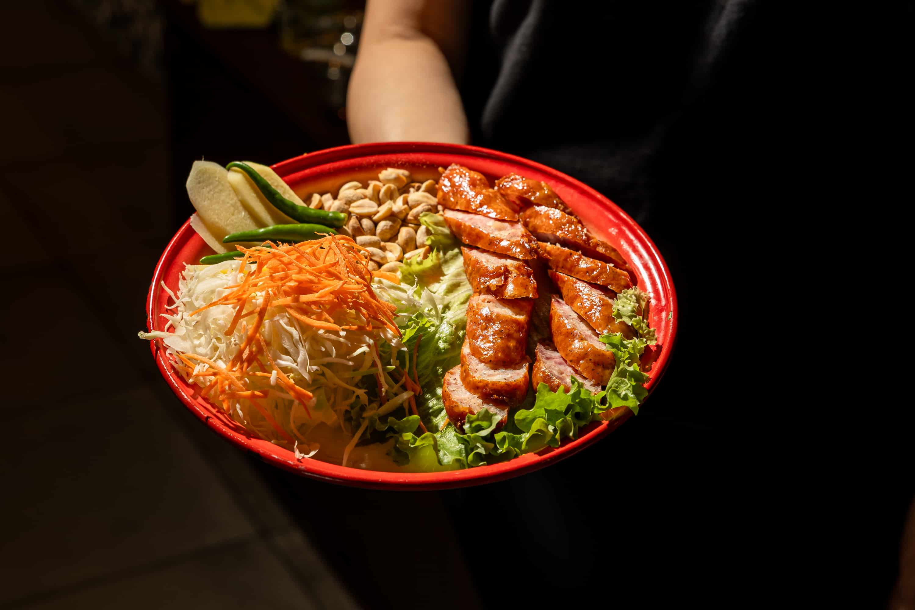 Red oval platter of sliced grilled sausage with shredded papaya, peanuts and lime, held aloft