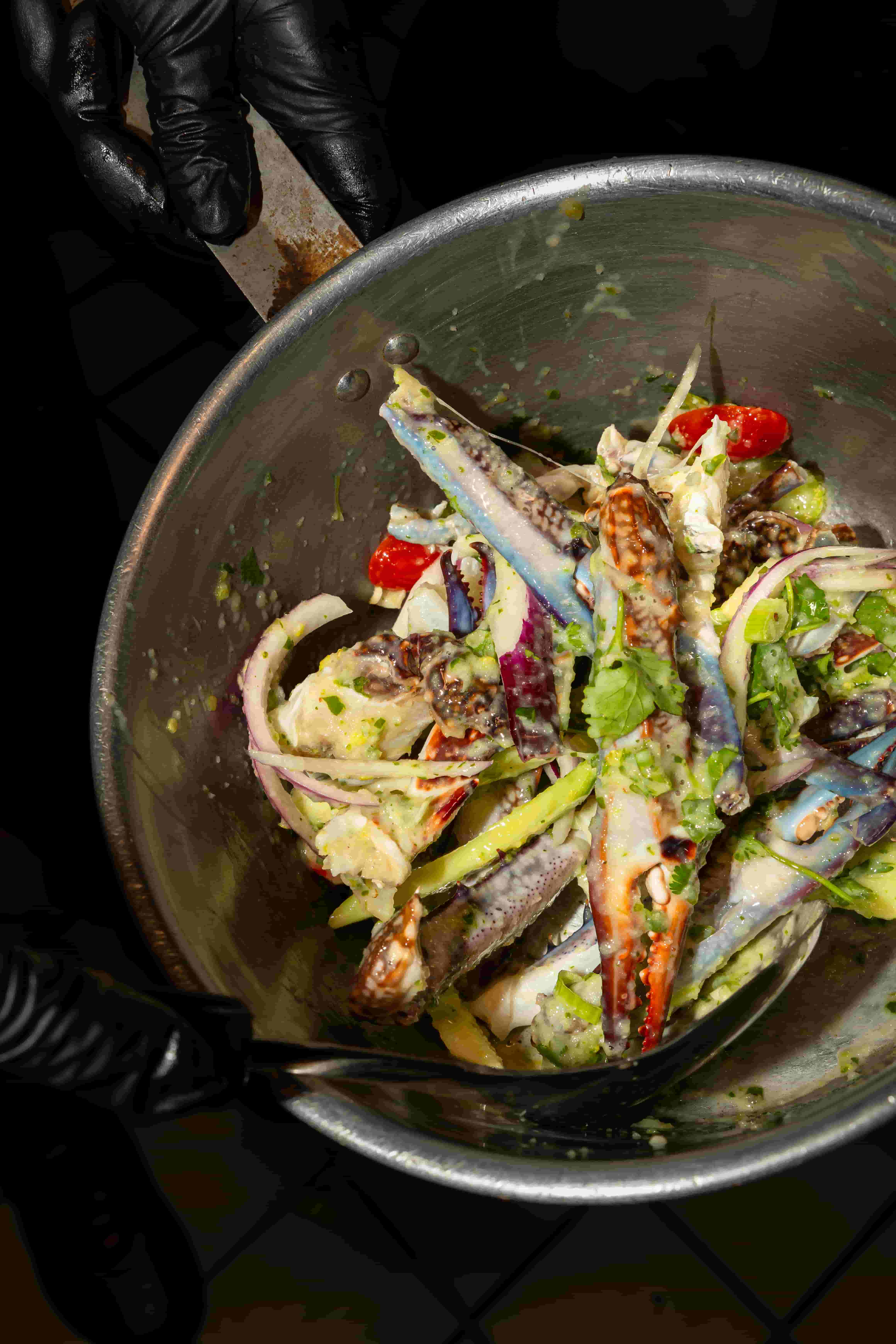 Gloved hands tossing blue crab pieces with cilantro, red onion and chilies in a metal bowl
