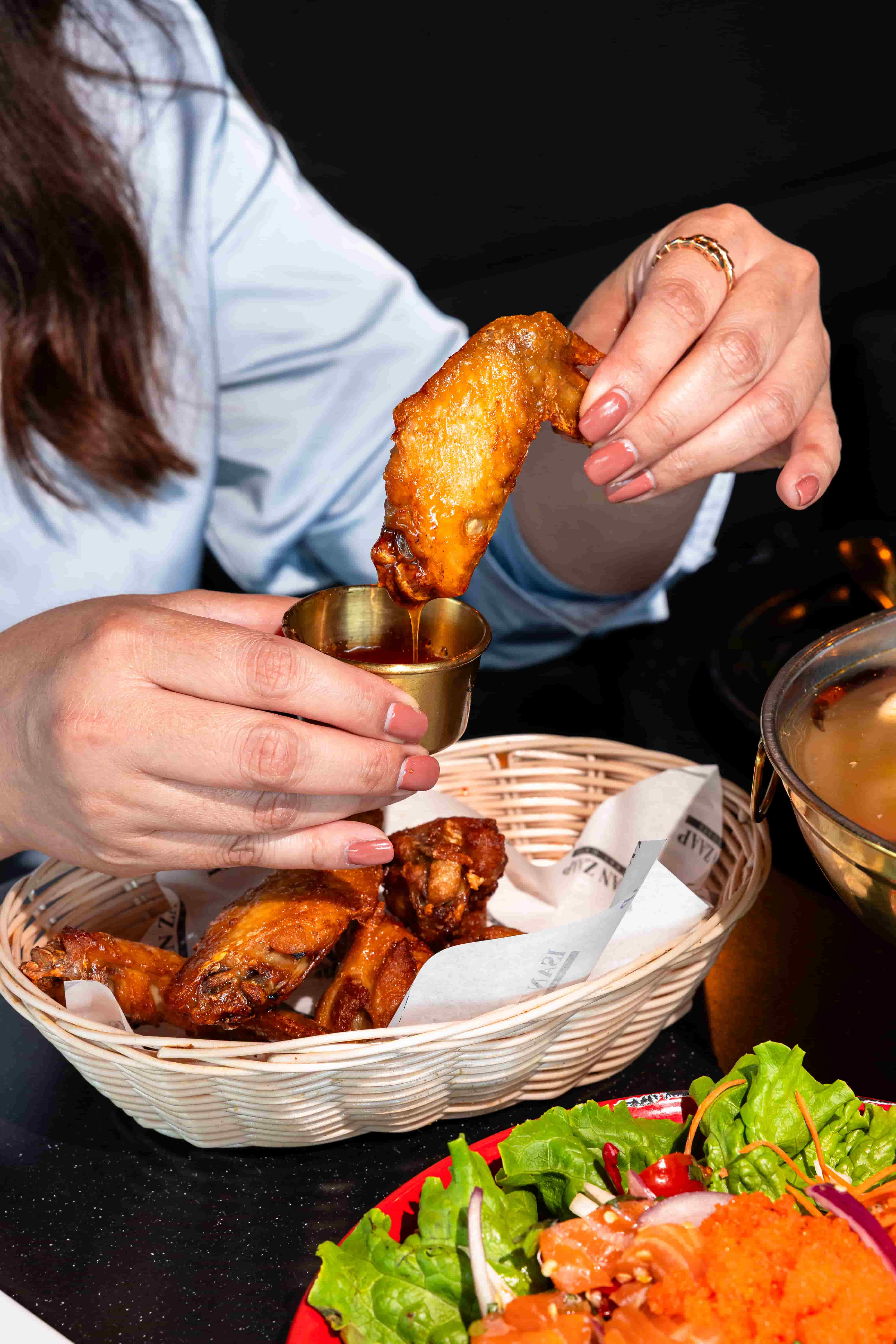 Fried chicken wing being dipped into a small gold cup of sauce above a wicker basket