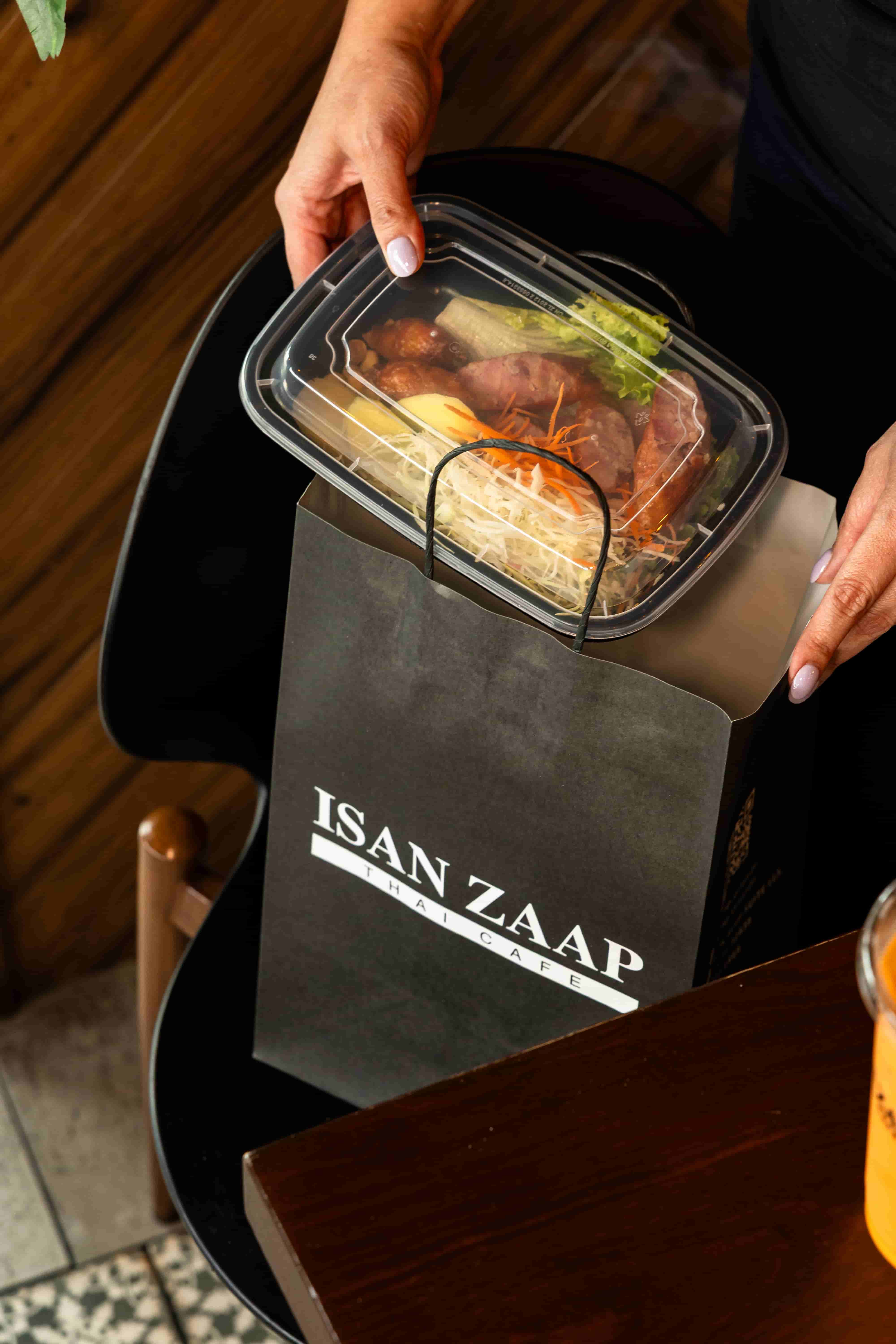 Clear takeout container of grilled meat and vegetables placed into a black Isan Zaap paper bag