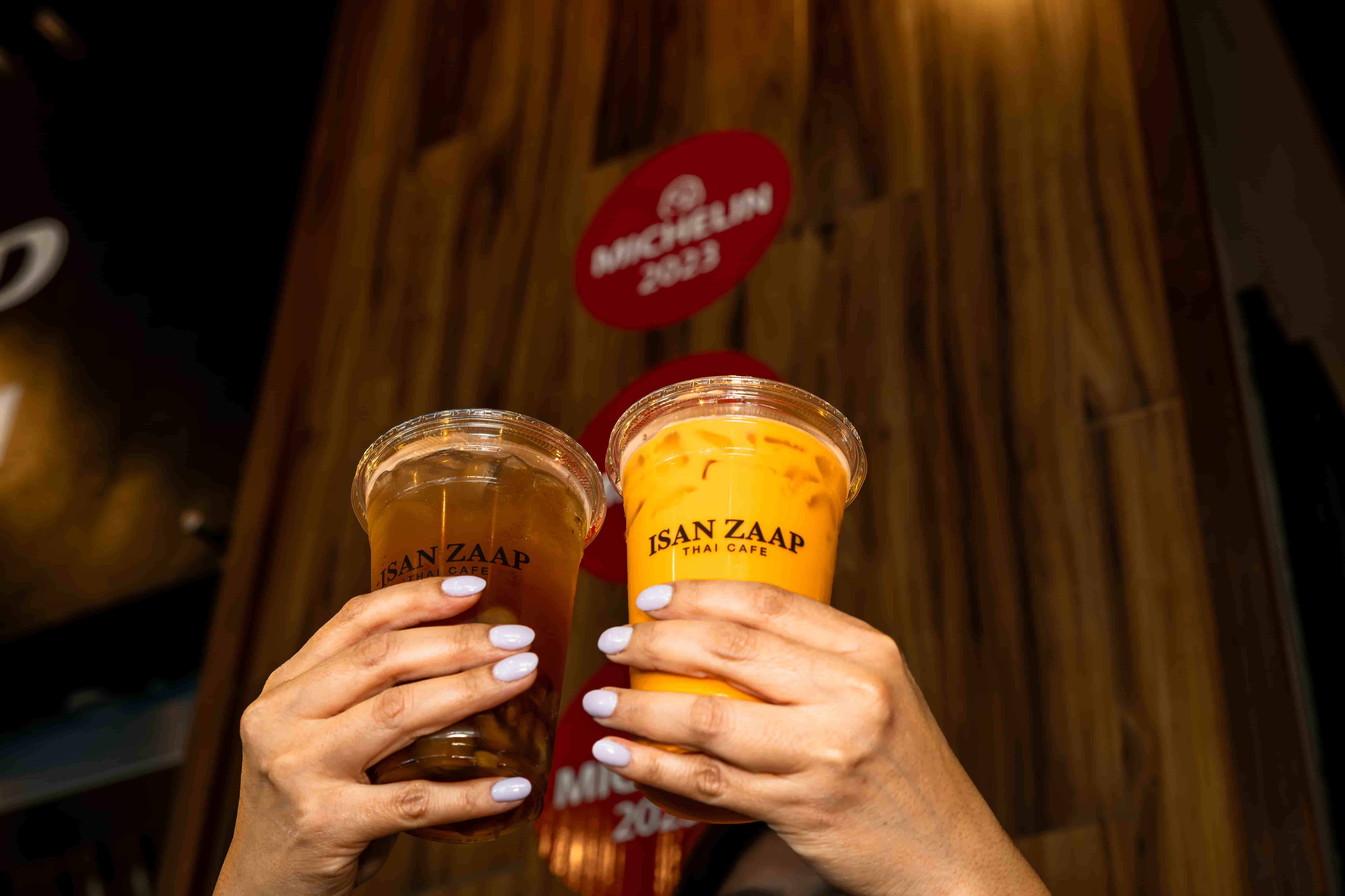 Two hands toasting branded cups of Thai iced tea in front of a MICHELIN 2023 plaque