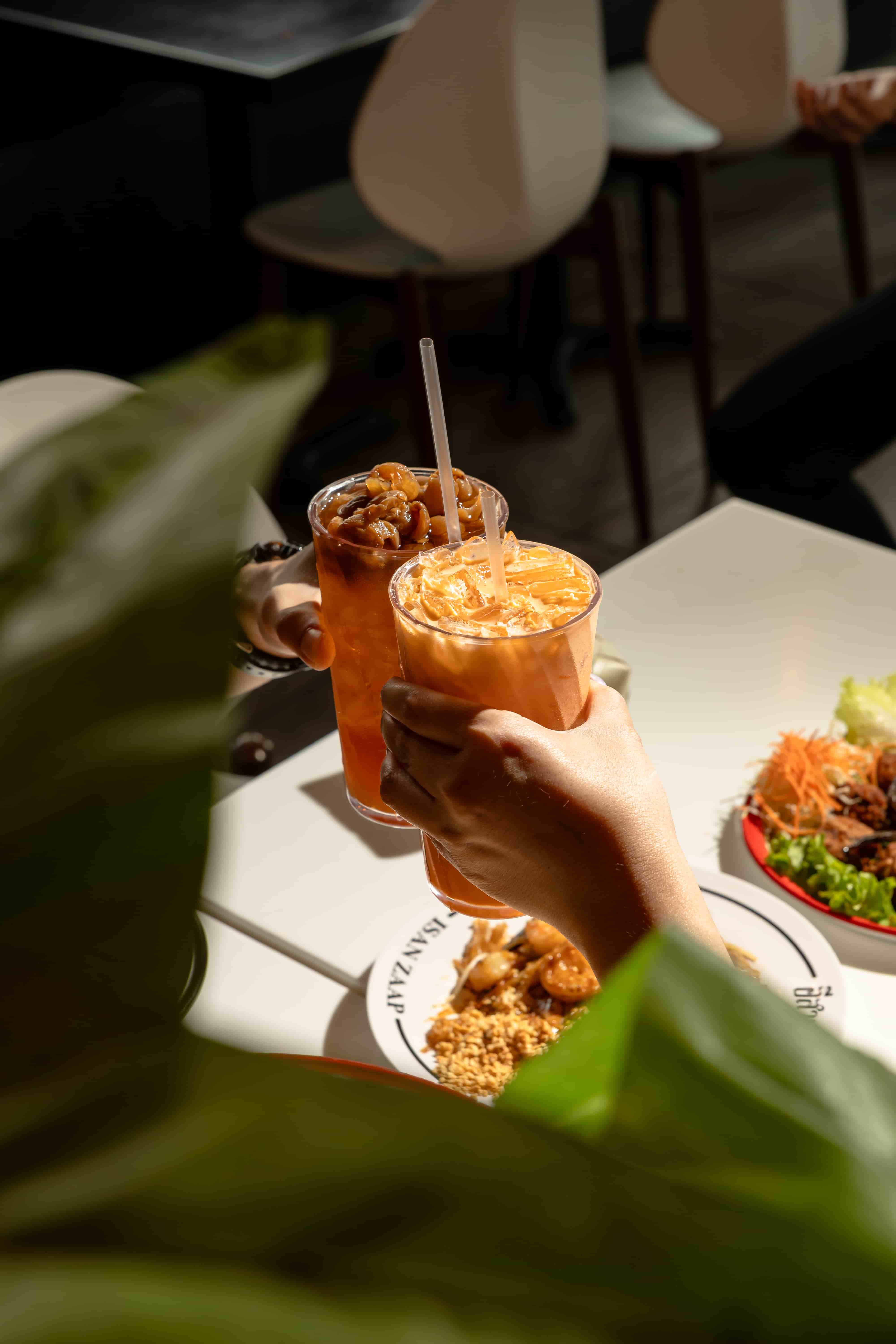 Two hands toasting glasses of Thai iced tea over a table of salad and crushed peanuts