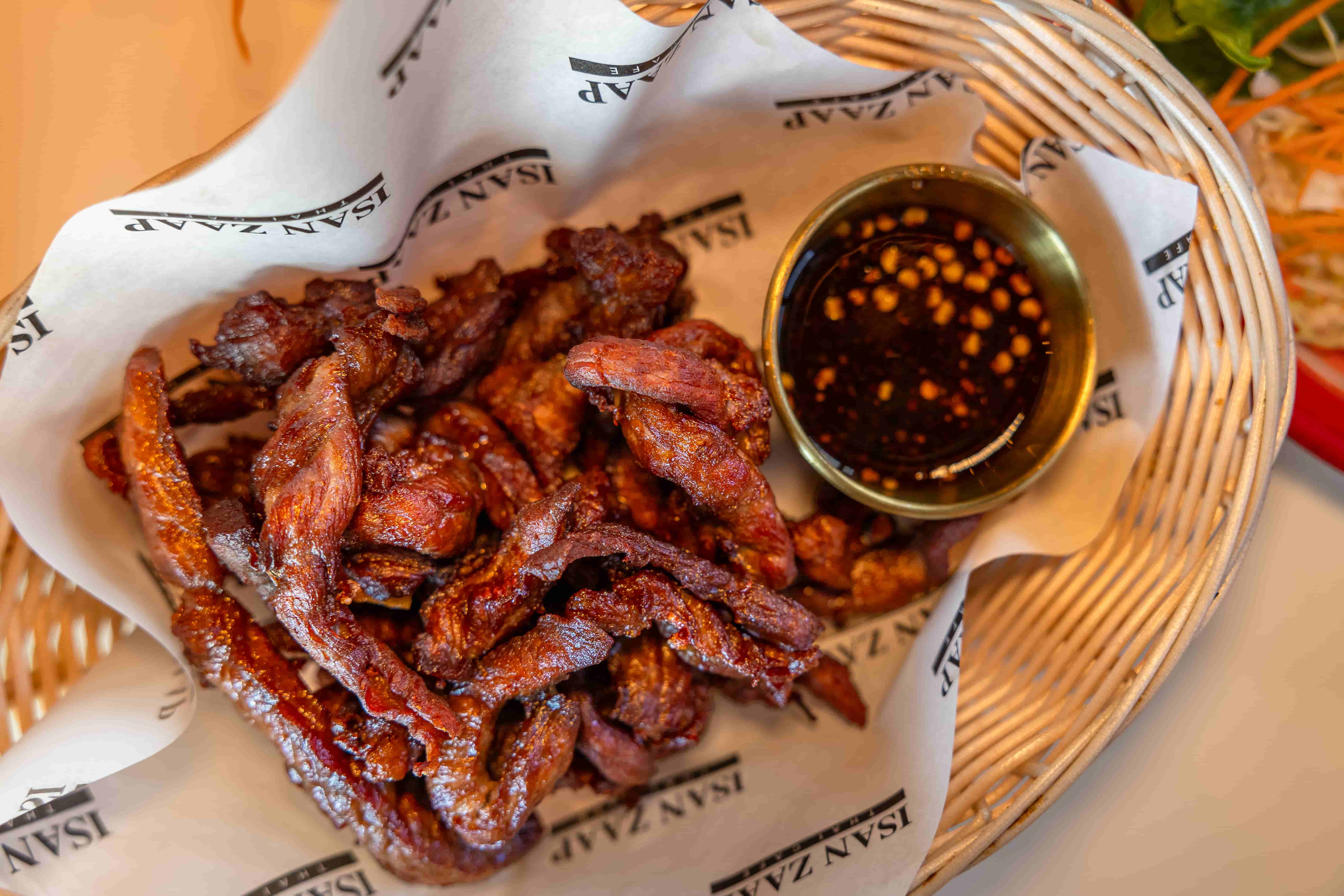 Basket of crisp fried pork strips lined with paper beside chili dipping sauce in a gold cup