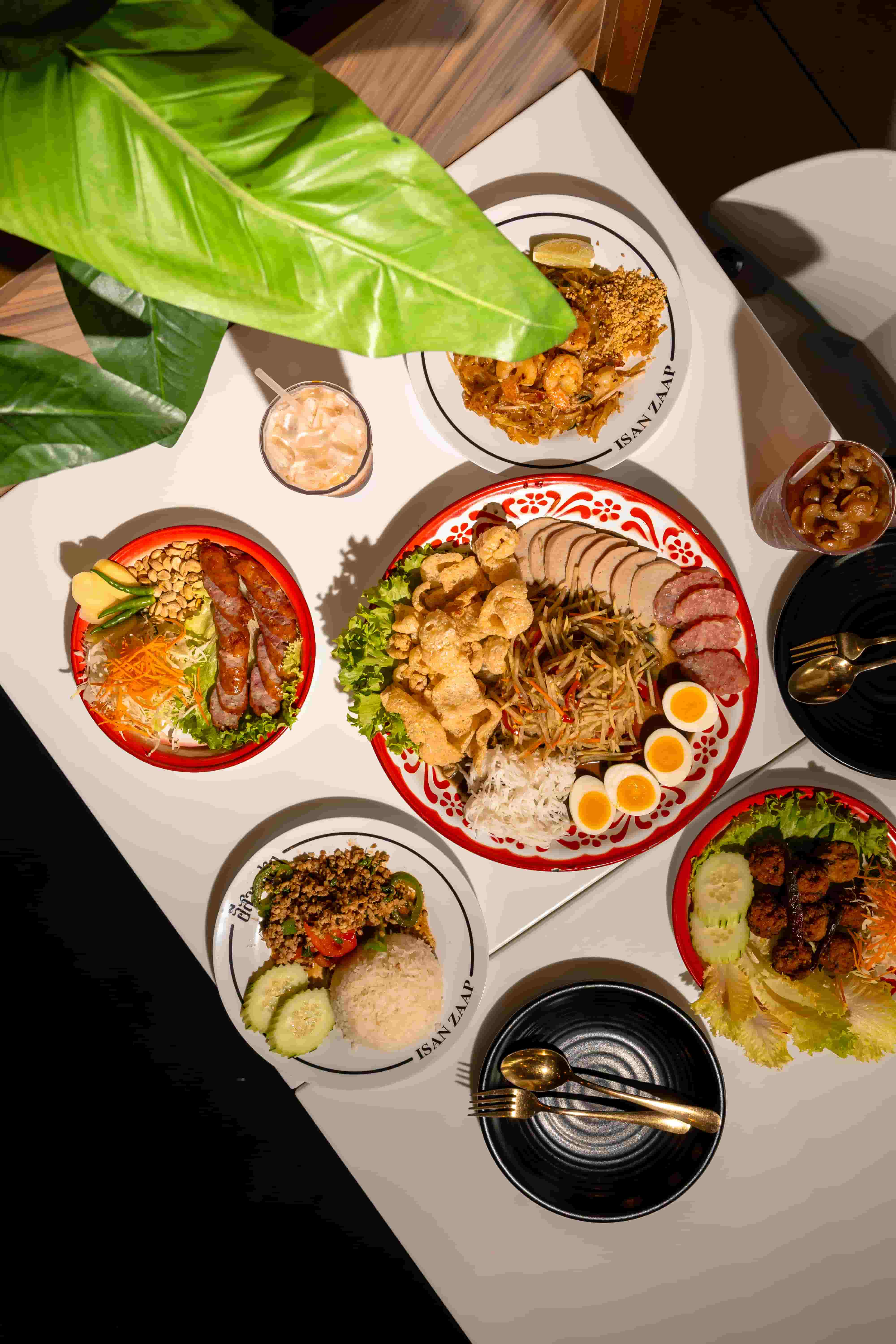 Overhead spread of papaya salad, grilled meats, sticky rice, noodles and boiled eggs on a white table