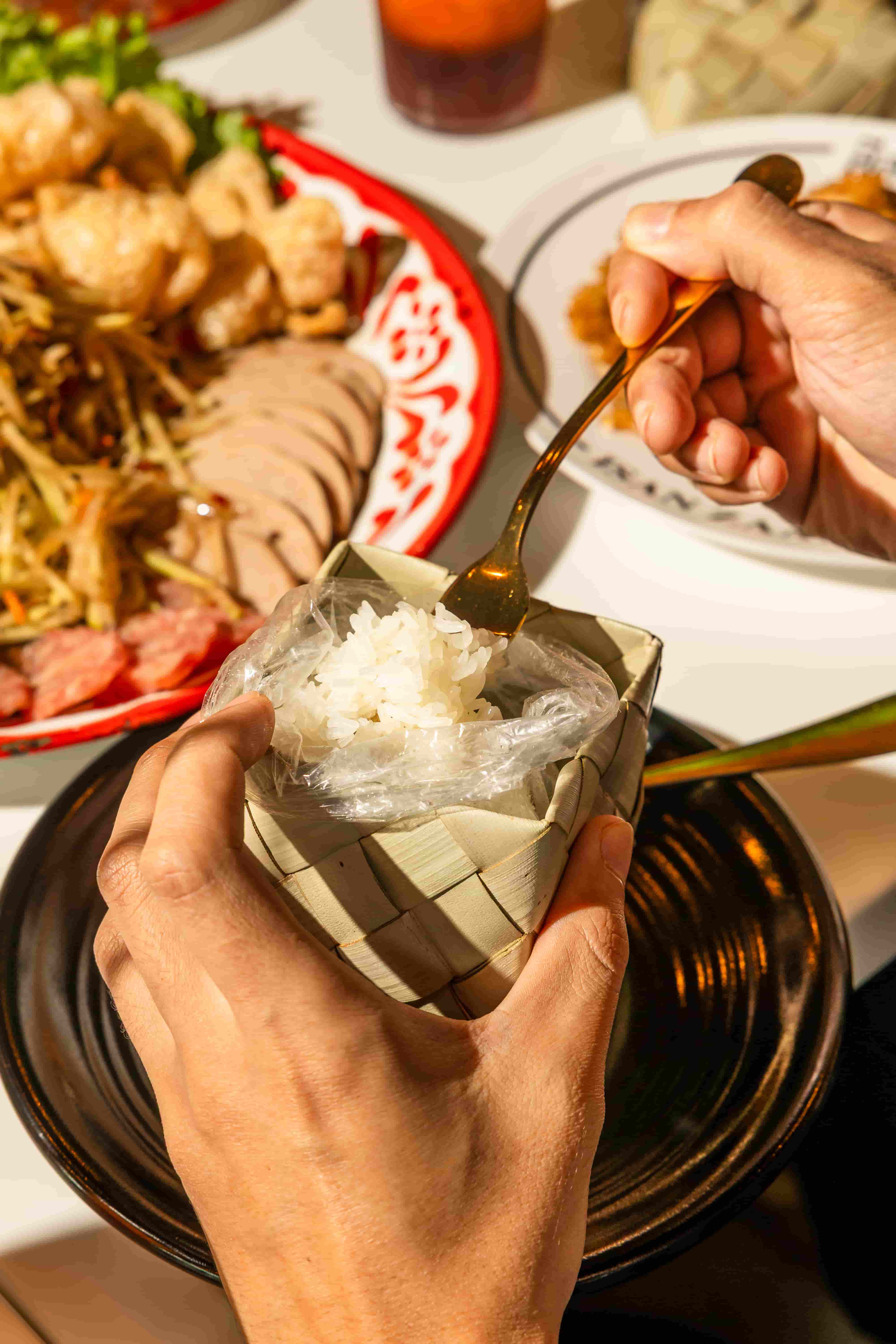 Hands holding a woven bamboo basket of sticky rice beside a red platter of grilled meats