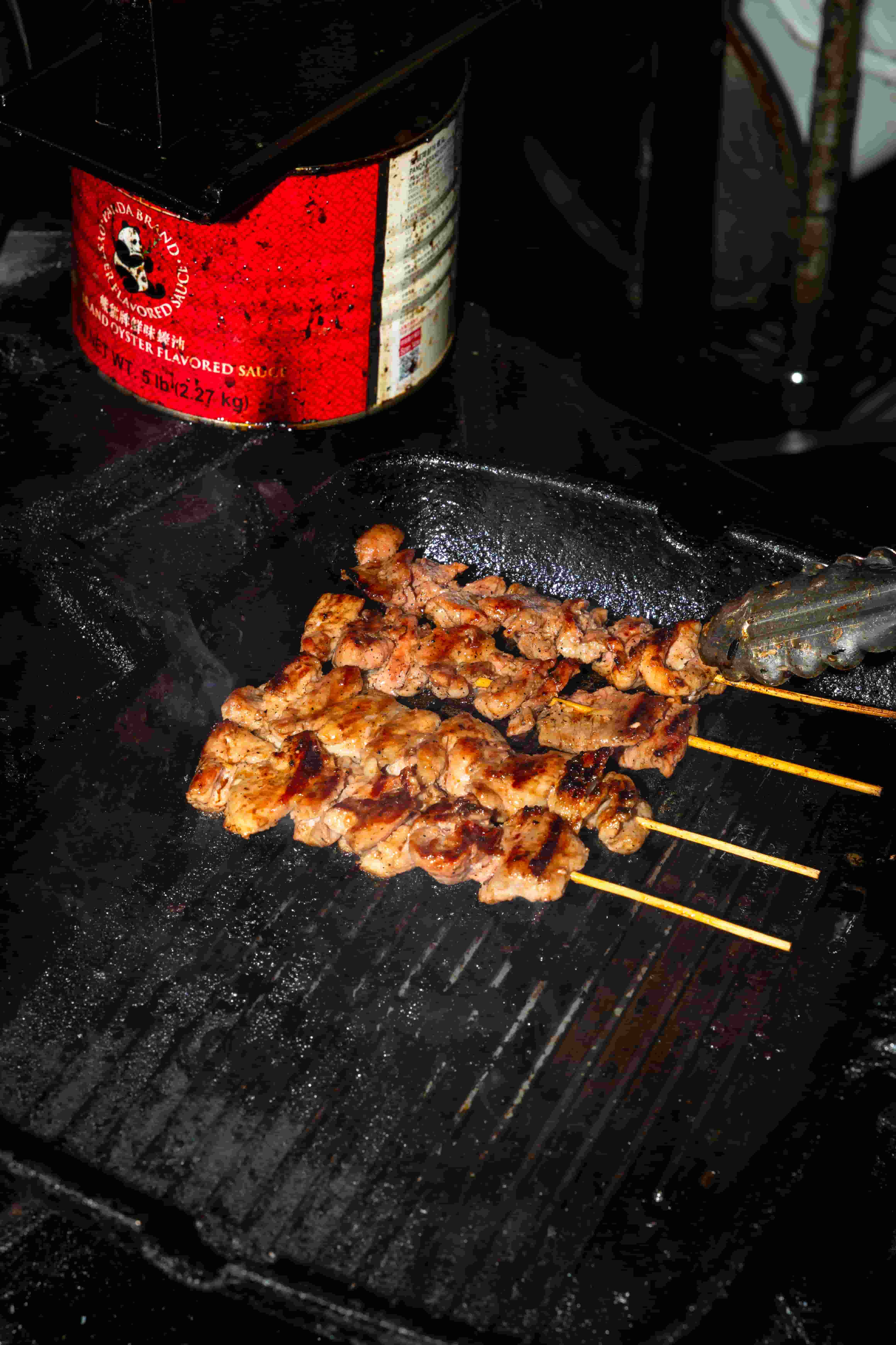 Pork skewers charring on a ribbed cast iron grill beside a tin of seasoning sauce