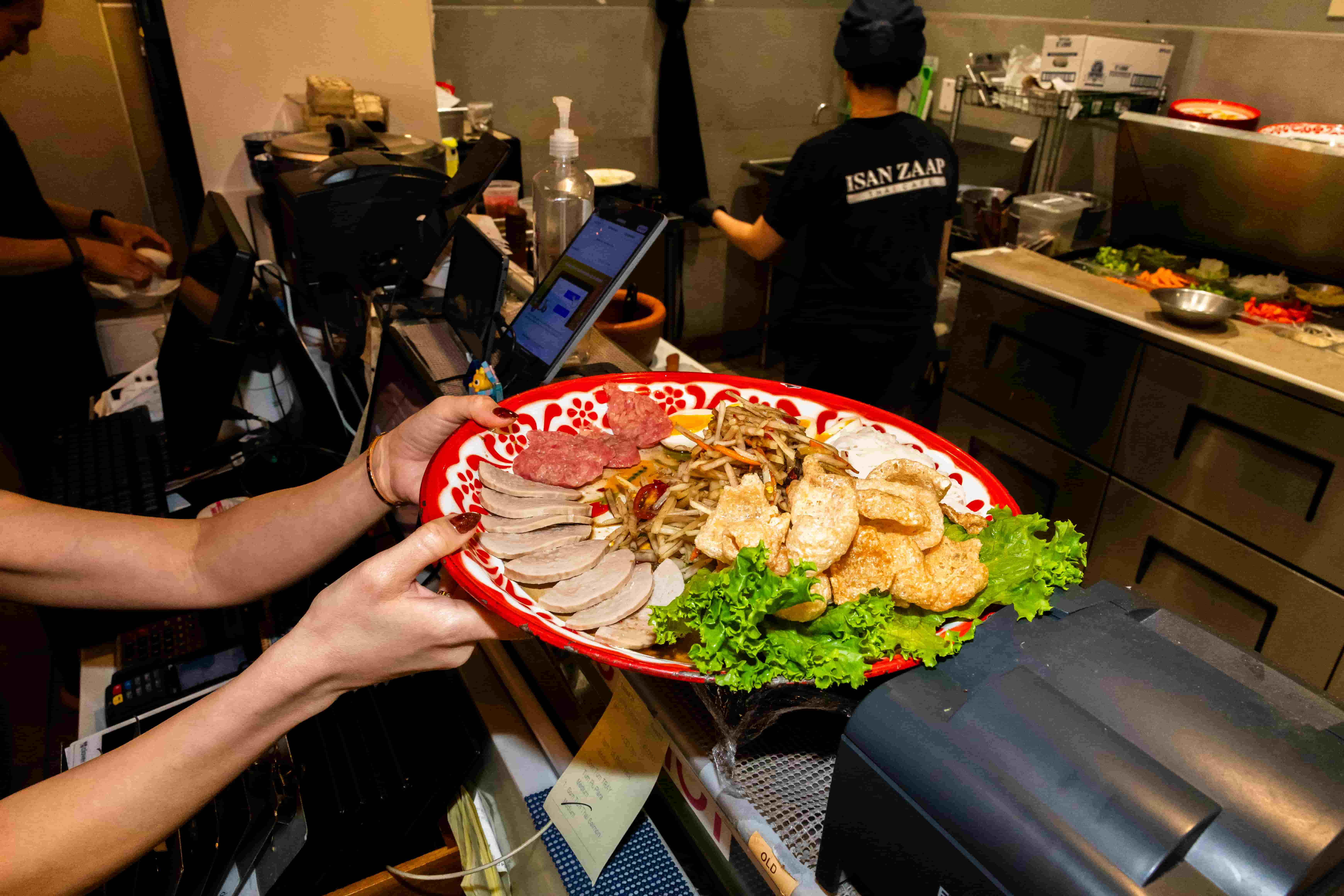 Server carrying a red platter of sliced sausage, pork rinds and lettuce through the open kitchen