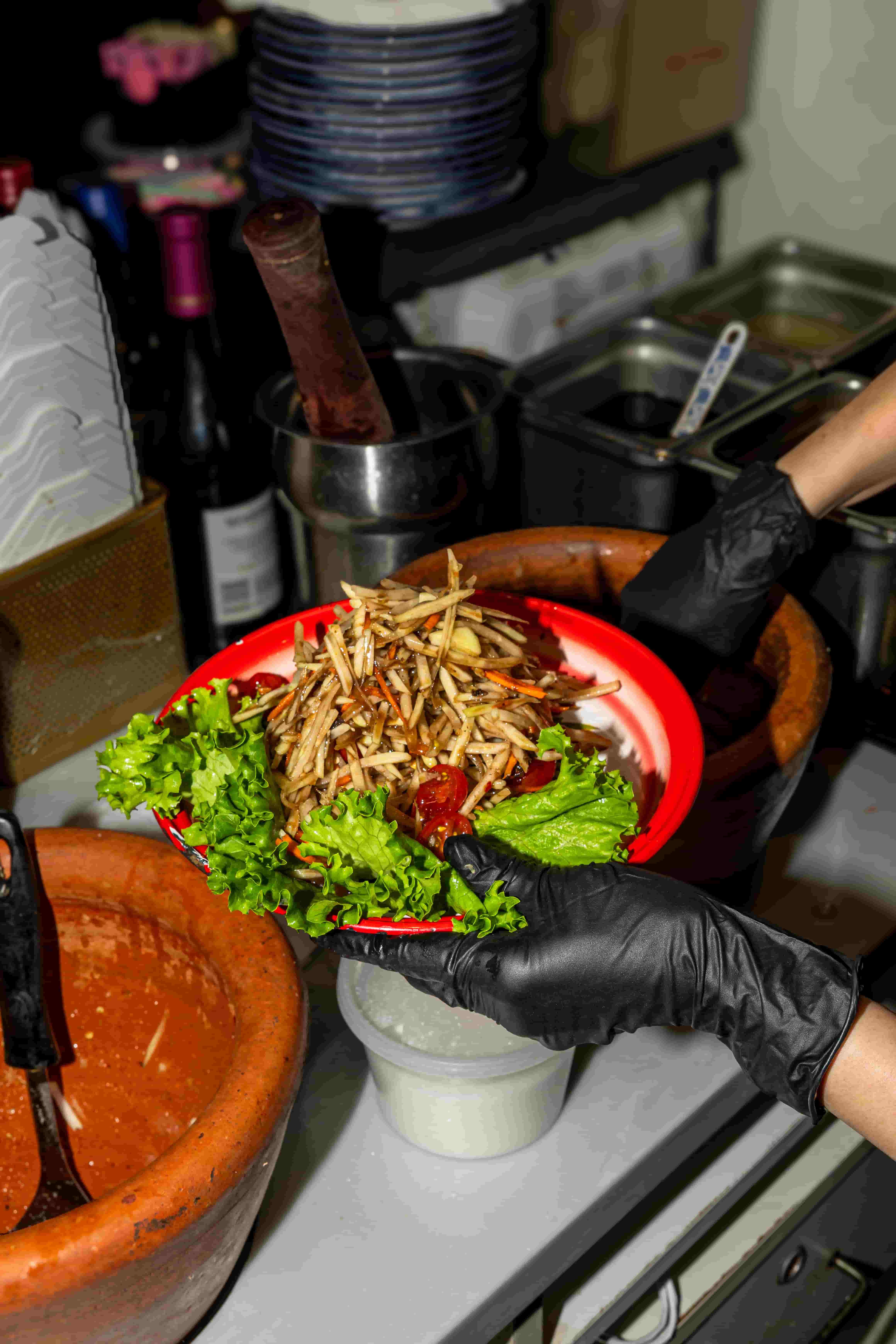Gloved hands plating shredded green papaya salad on a red plate beside a clay mortar