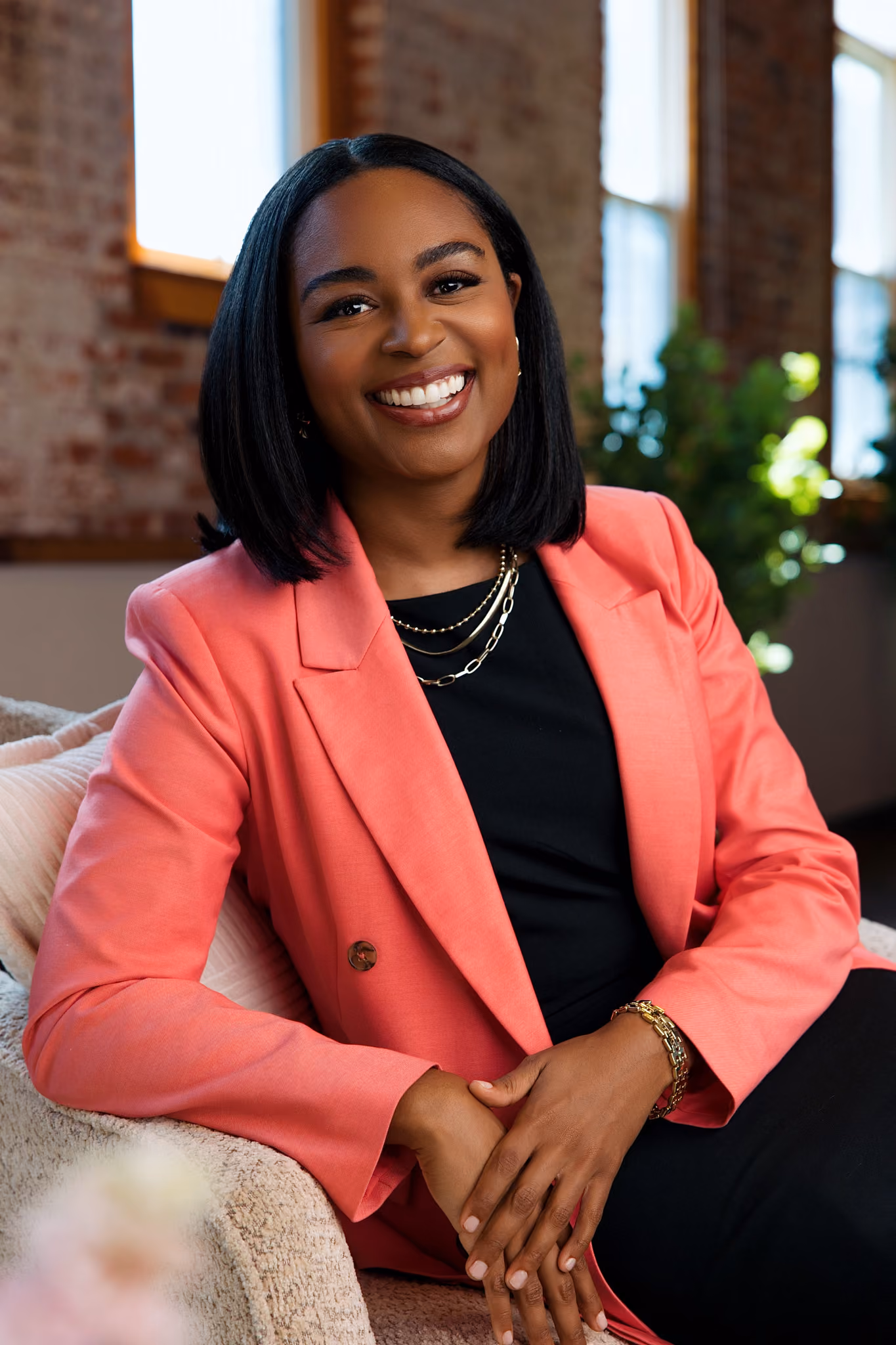 Smiling woman with shoulder-length black hair wearing a coral blazer, black top, and gold jewelry, seated on a beige chair in a room with brick walls and windows.