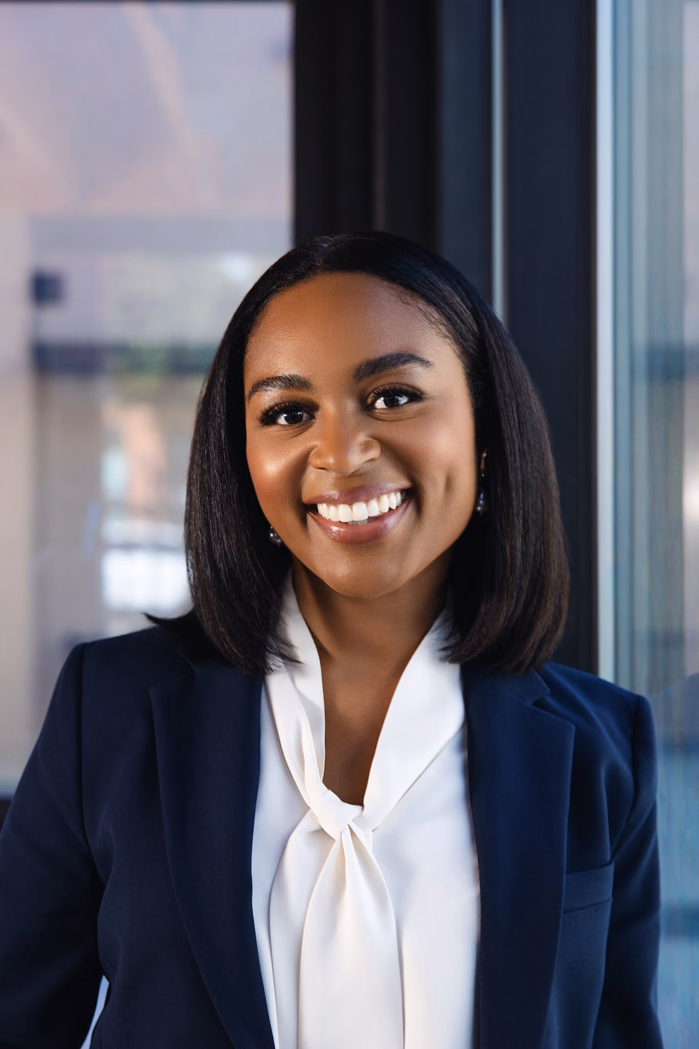 Smiling professional Black woman with straight shoulder-length hair wearing a dark blazer and white blouse, standing indoors near a window.