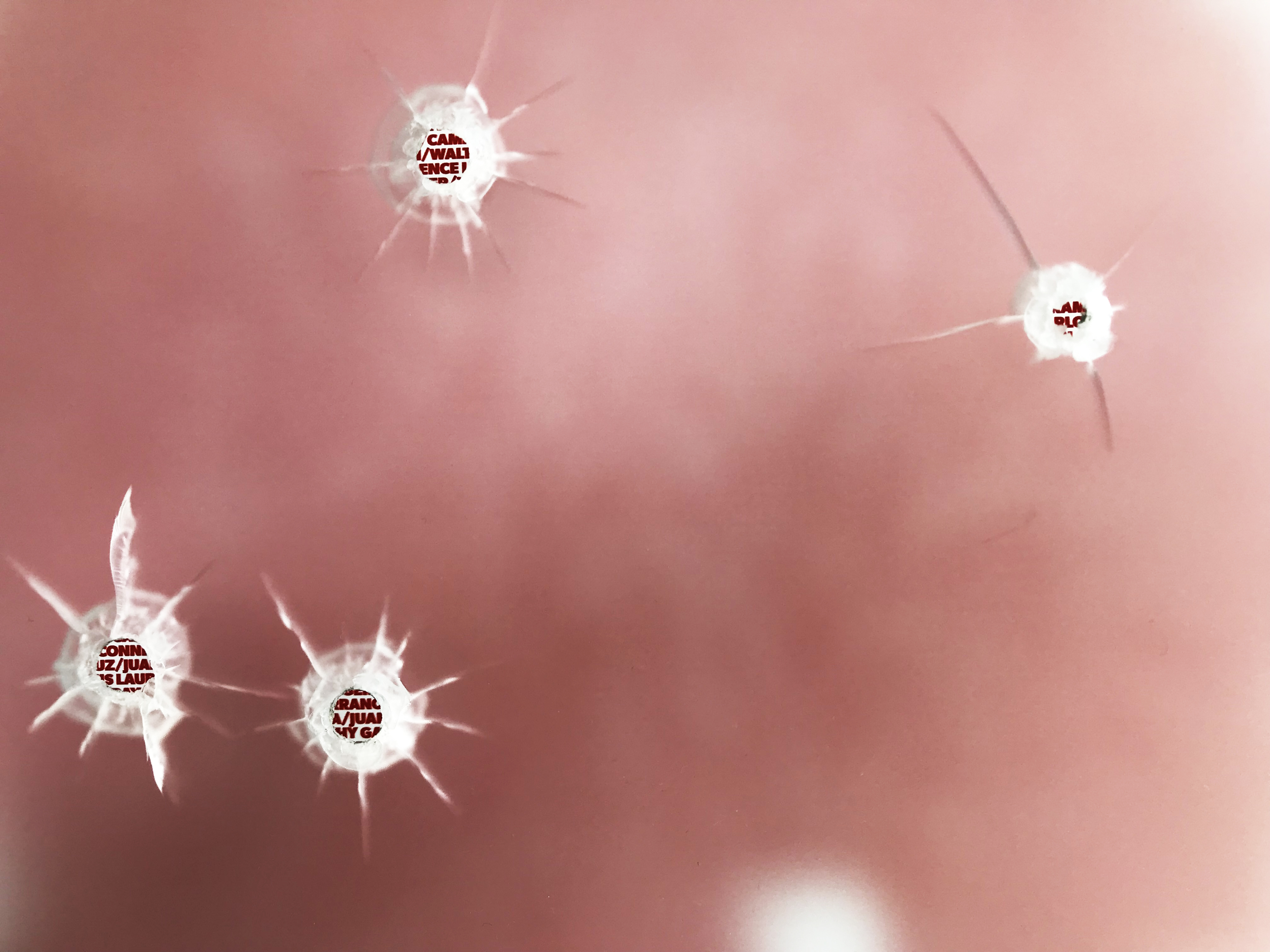 Four bullet holes in the frosted acrylic, cracks radiating from each, over a blur of red.